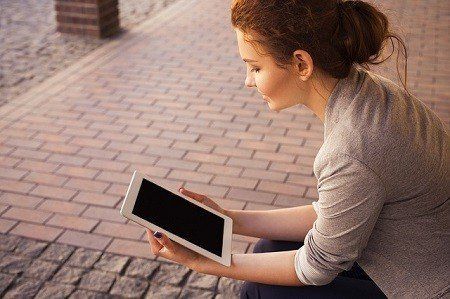 A woman is sitting on the sidewalk using a tablet computer.